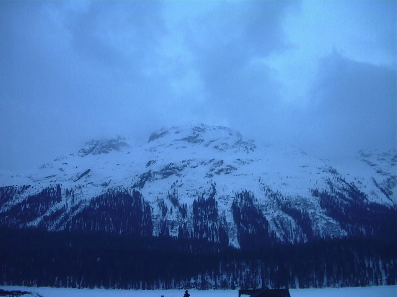 Blue-toned clouds hang over a snowy mountain above a dark forest.