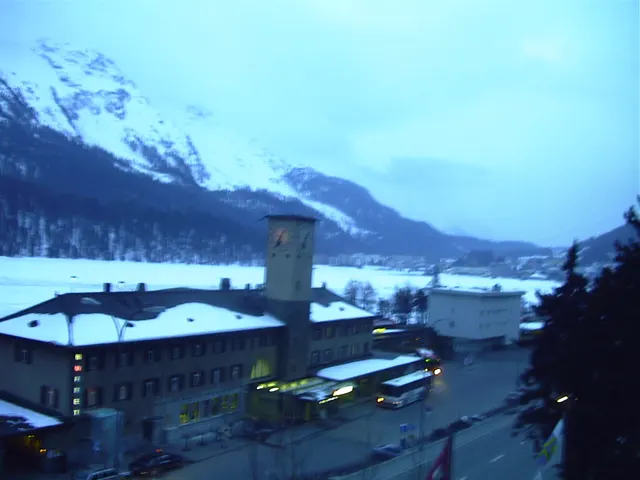 Lights glow around a snow-covered station building beneath blue winter mountains.
