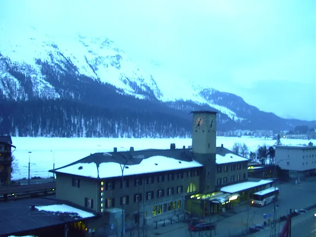 A station building and its tower stand below snowy mountains in cool evening light.