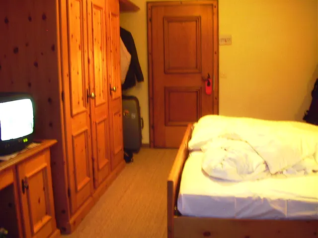 A pale bed stands beside tall wooden cupboards and a television in a warmly lit room.