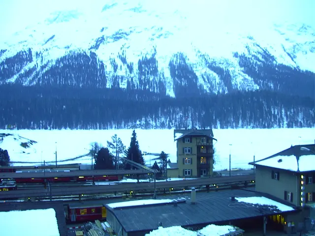 Tracks, a small tower and snowy roofs lie beneath a broad white mountain ridge.
