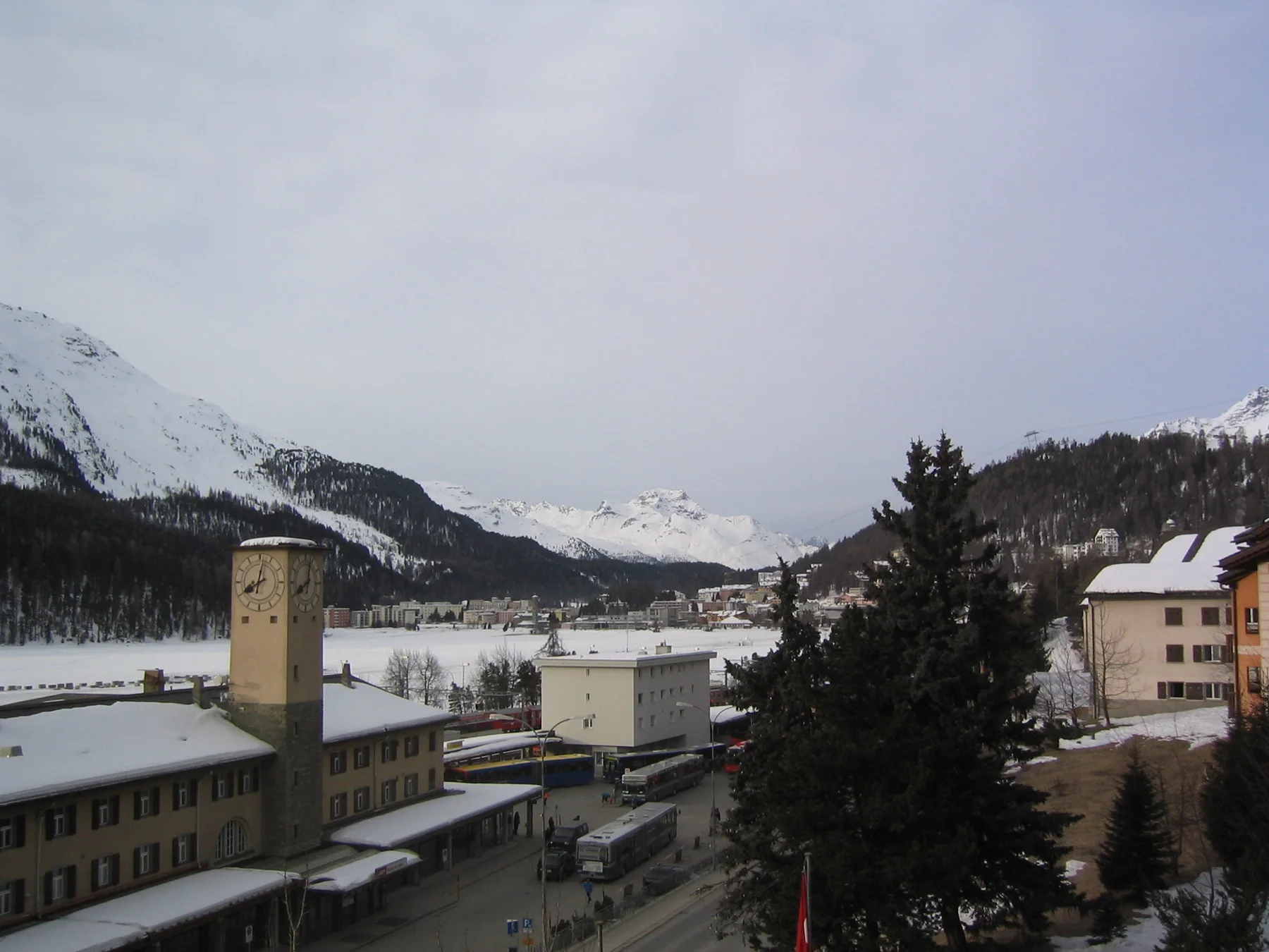 A station tower and roadside buildings stand beside snow beneath an overcast mountain sky.