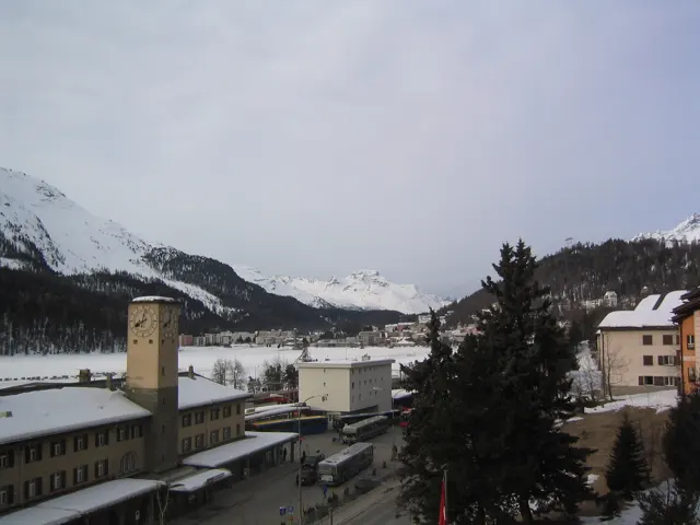 A station tower and roadside buildings stand beside snow beneath an overcast mountain sky.