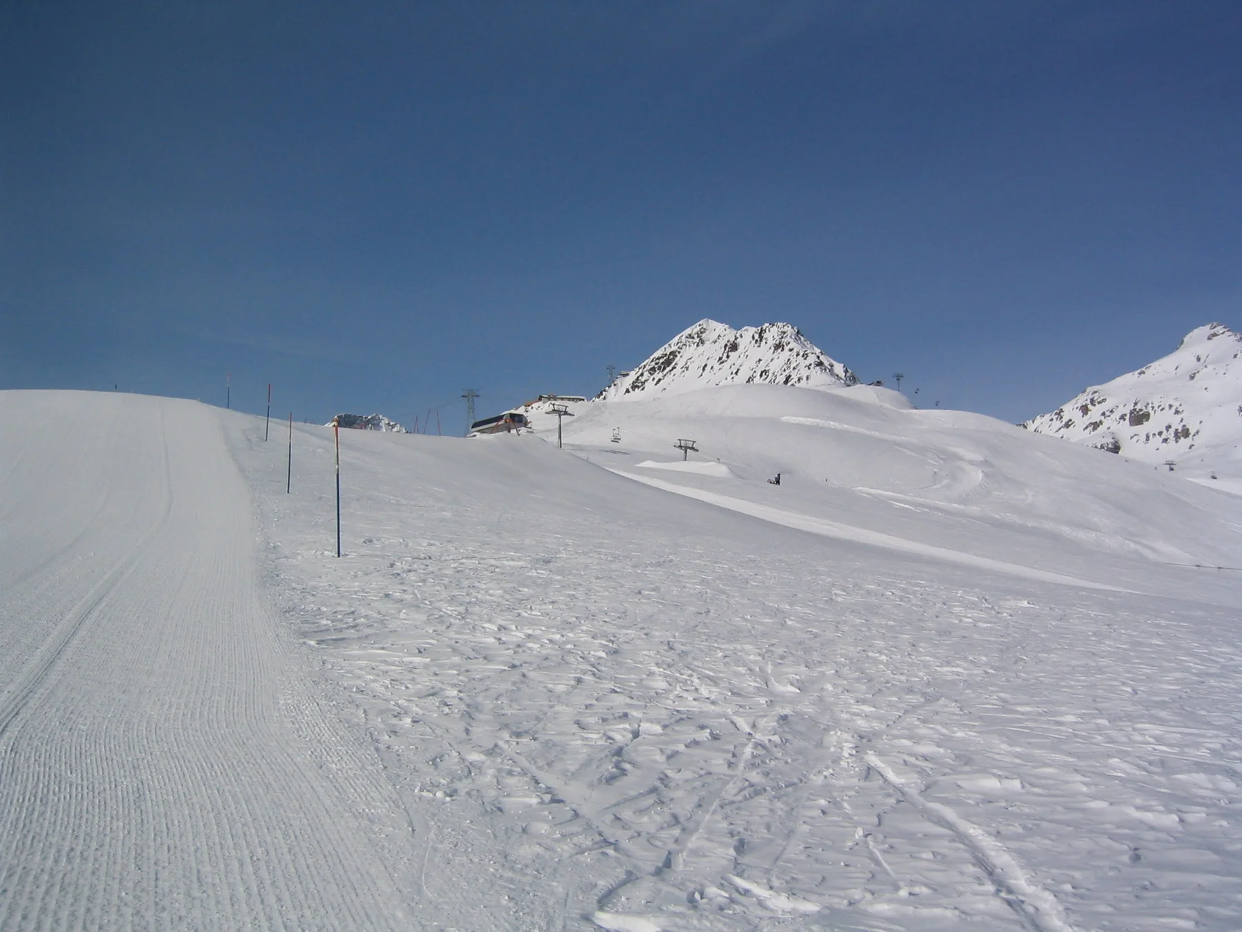 A marked snowy track leads toward a pointed white peak under blue sky.