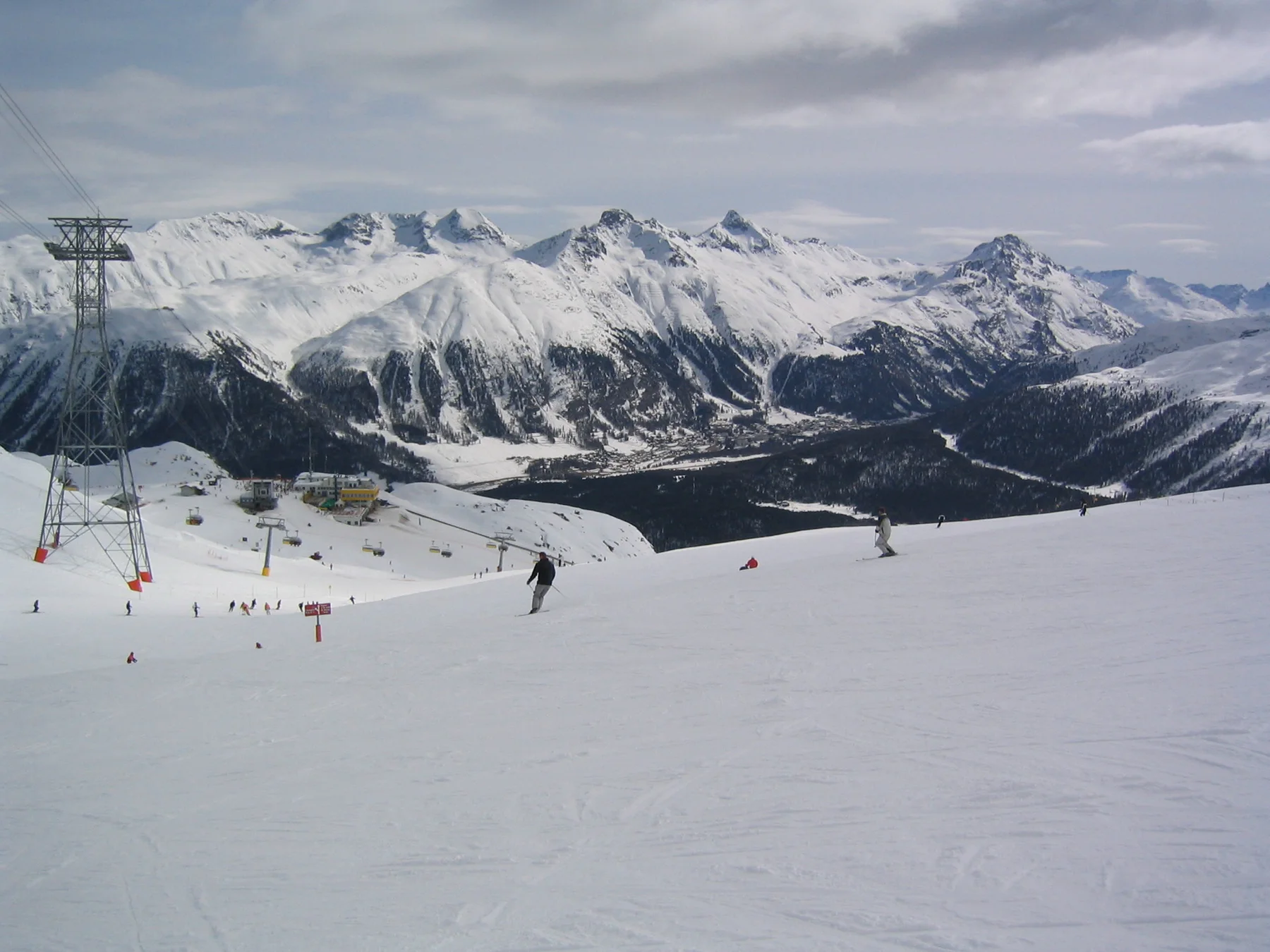 Scattered skiers cross a wide slope overlooking a deep mountain valley.
