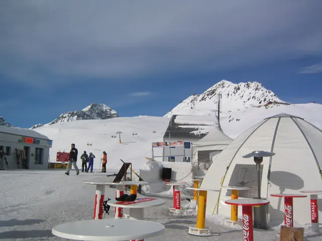 Outdoor tables and a pale dome-shaped structure stand before a snowy mountain peak.