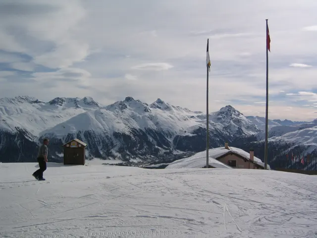 Tall flagpoles stand on a snow-covered terrace overlooking distant mountain ranges.