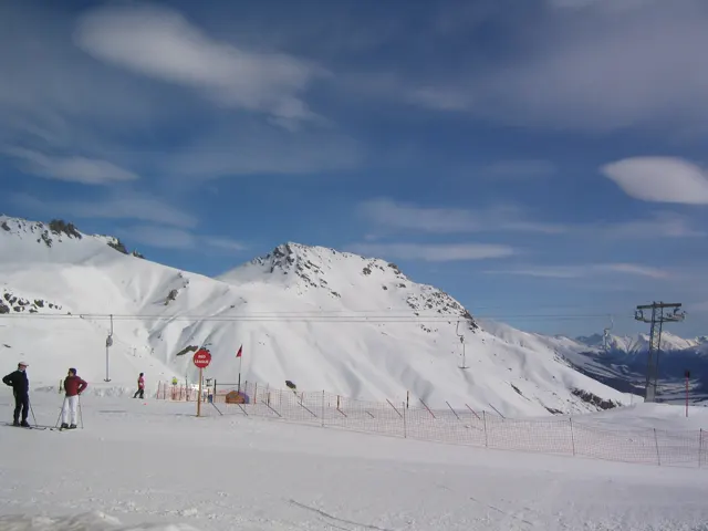Two people stand on a snowy plateau with mountain ridges and lift structures beyond.