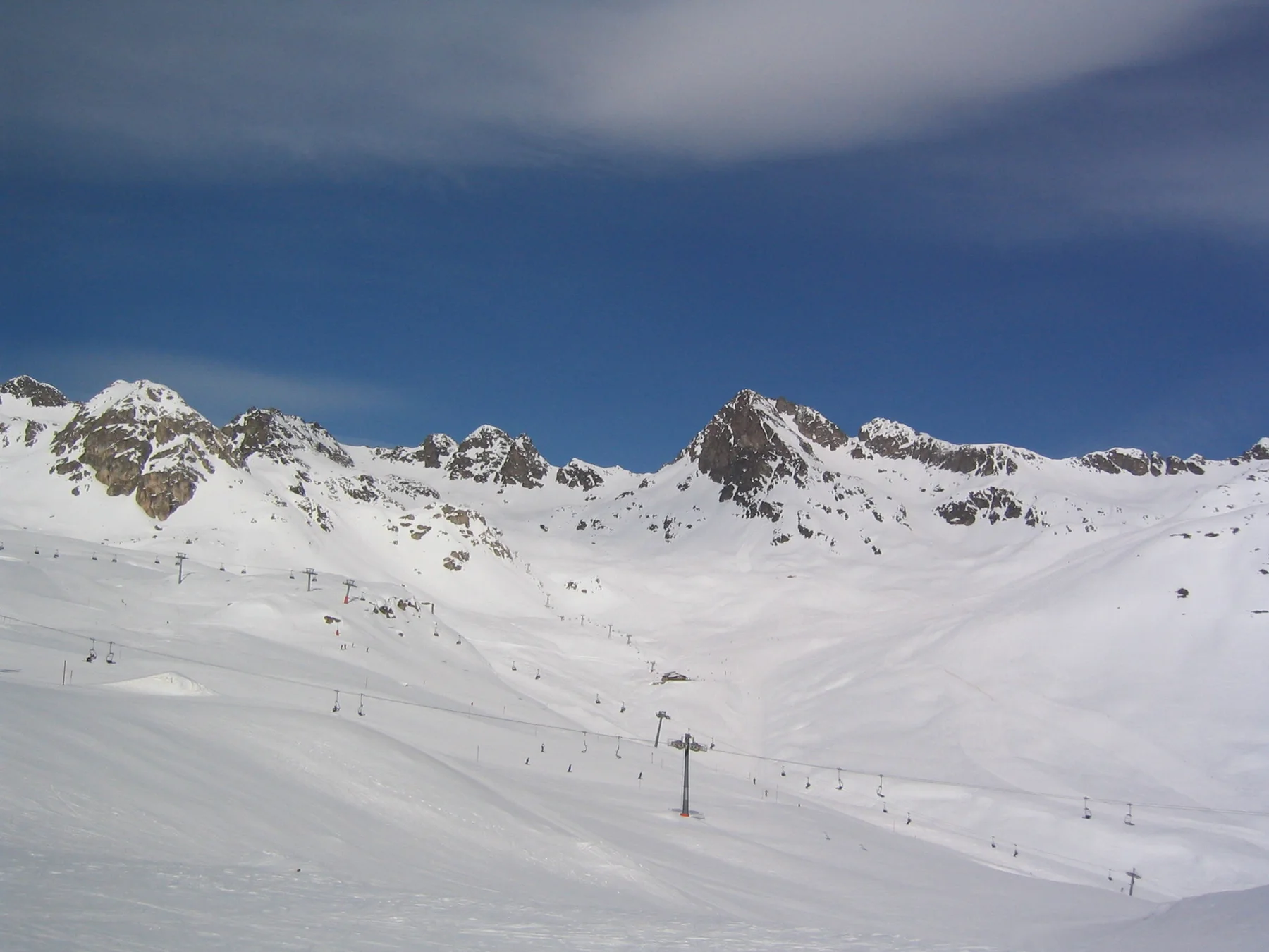 Jagged rocky peaks line a broad white basin crossed by ski lifts.