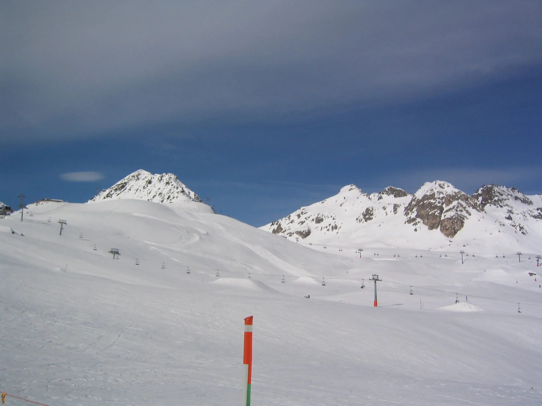 An orange piste marker stands before rolling snow slopes and rocky peaks.