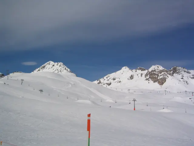An orange piste marker stands before rolling snow slopes and rocky peaks.