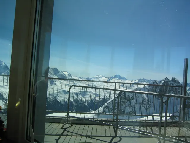 A window and balcony railing frame a high panorama of snowy mountains.