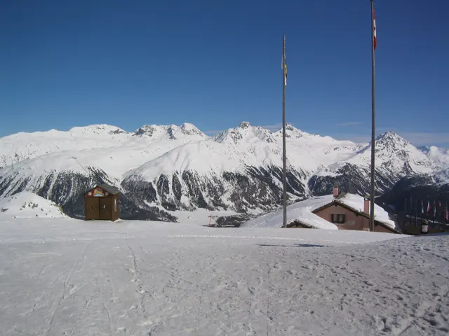 Tall flagpoles and small roofed structures stand on a snowy terrace above distant peaks.