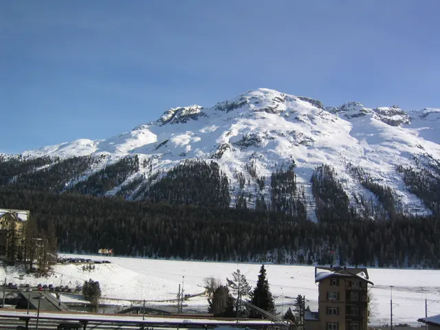 A bright snowy mountain rises above a dark forest and railway tracks.