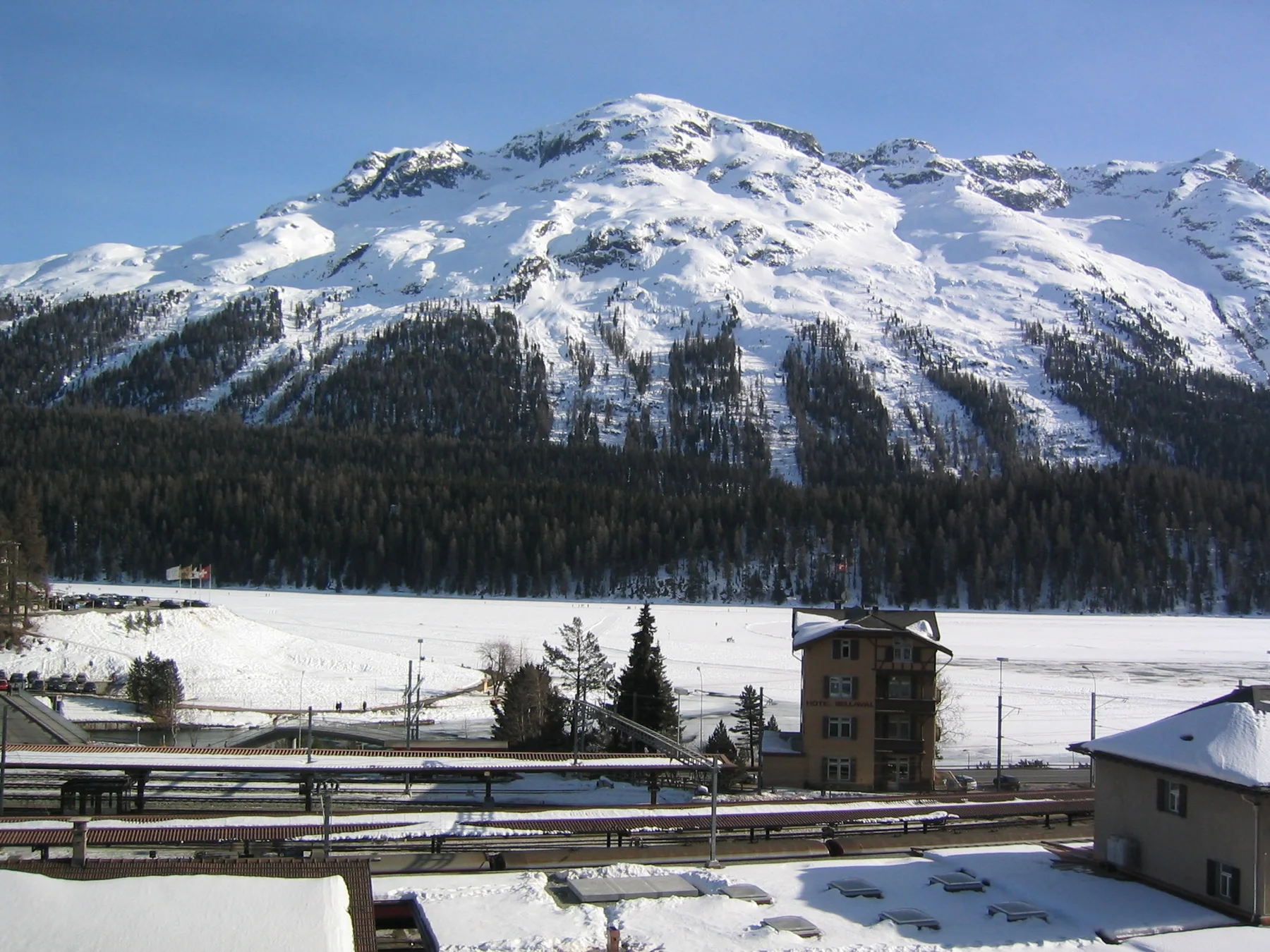 Railway tracks and a small tower cross the snowy foreground below a sunlit mountain.