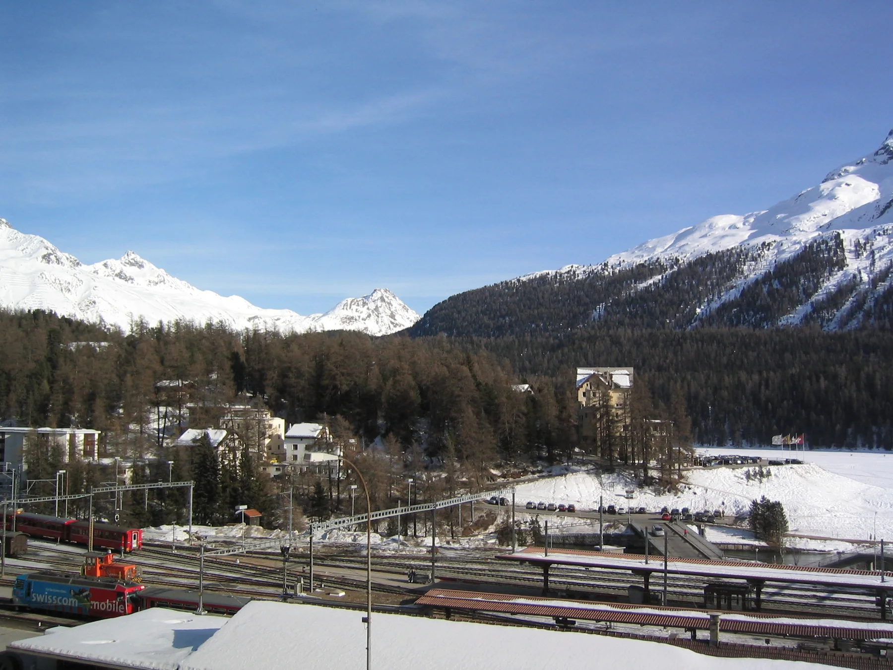 A railway yard fills the foreground of a snow-covered valley beneath distant mountains.