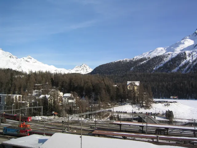 A railway yard fills the foreground of a snow-covered valley beneath distant mountains.