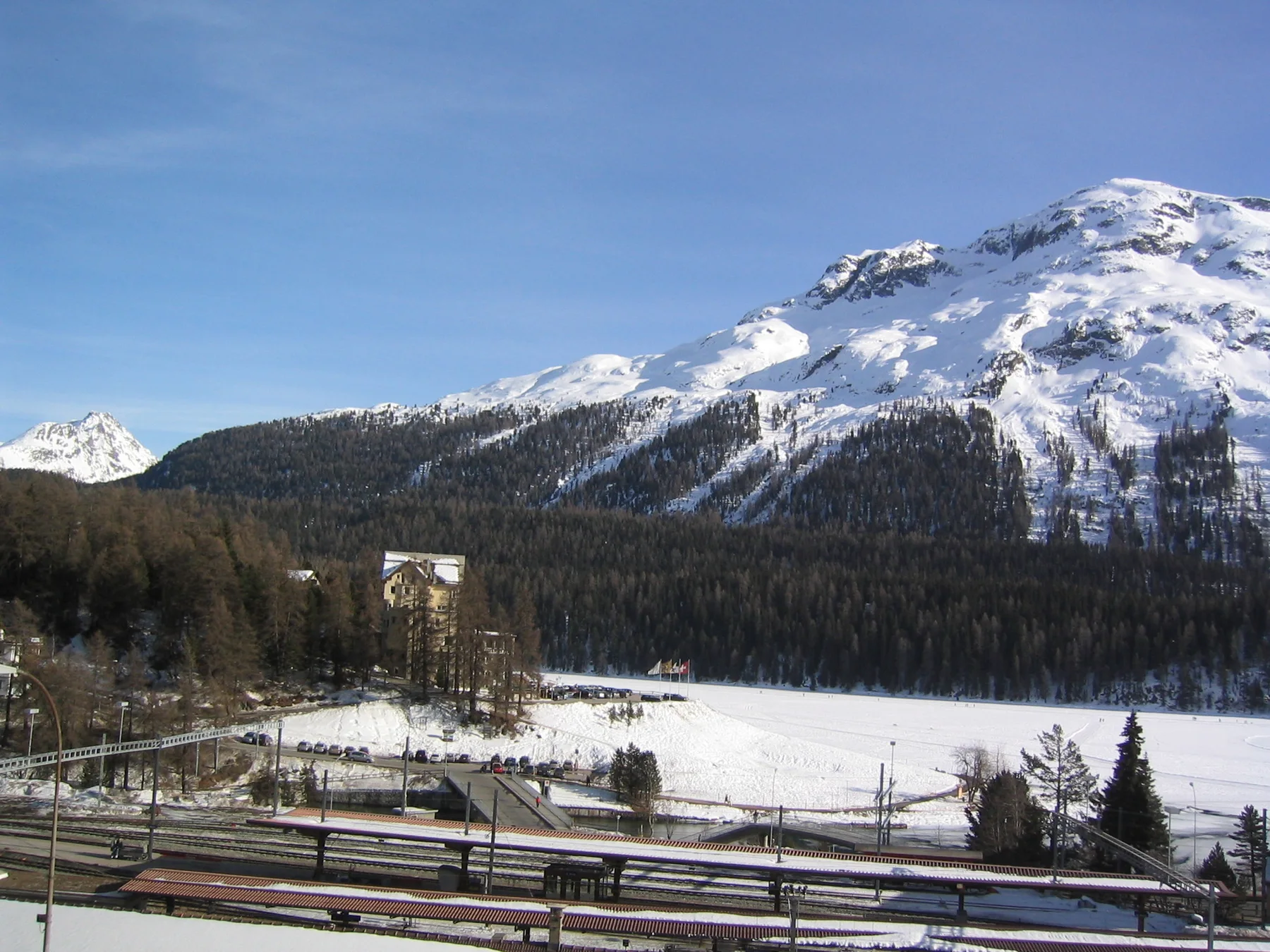 Tracks and overhead wires lead across a winter valley below a broad snowy ridge.