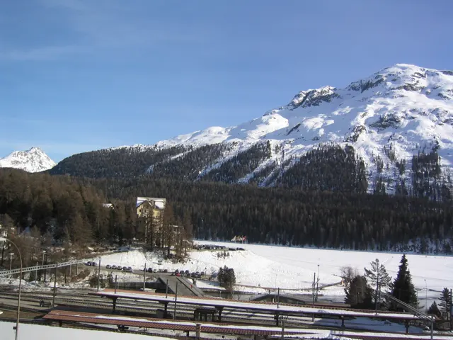 Tracks and overhead wires lead across a winter valley below a broad snowy ridge.