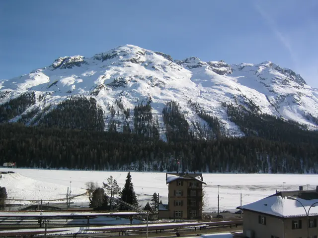 A small tower stands among snowy railway grounds beneath a sunlit mountain.