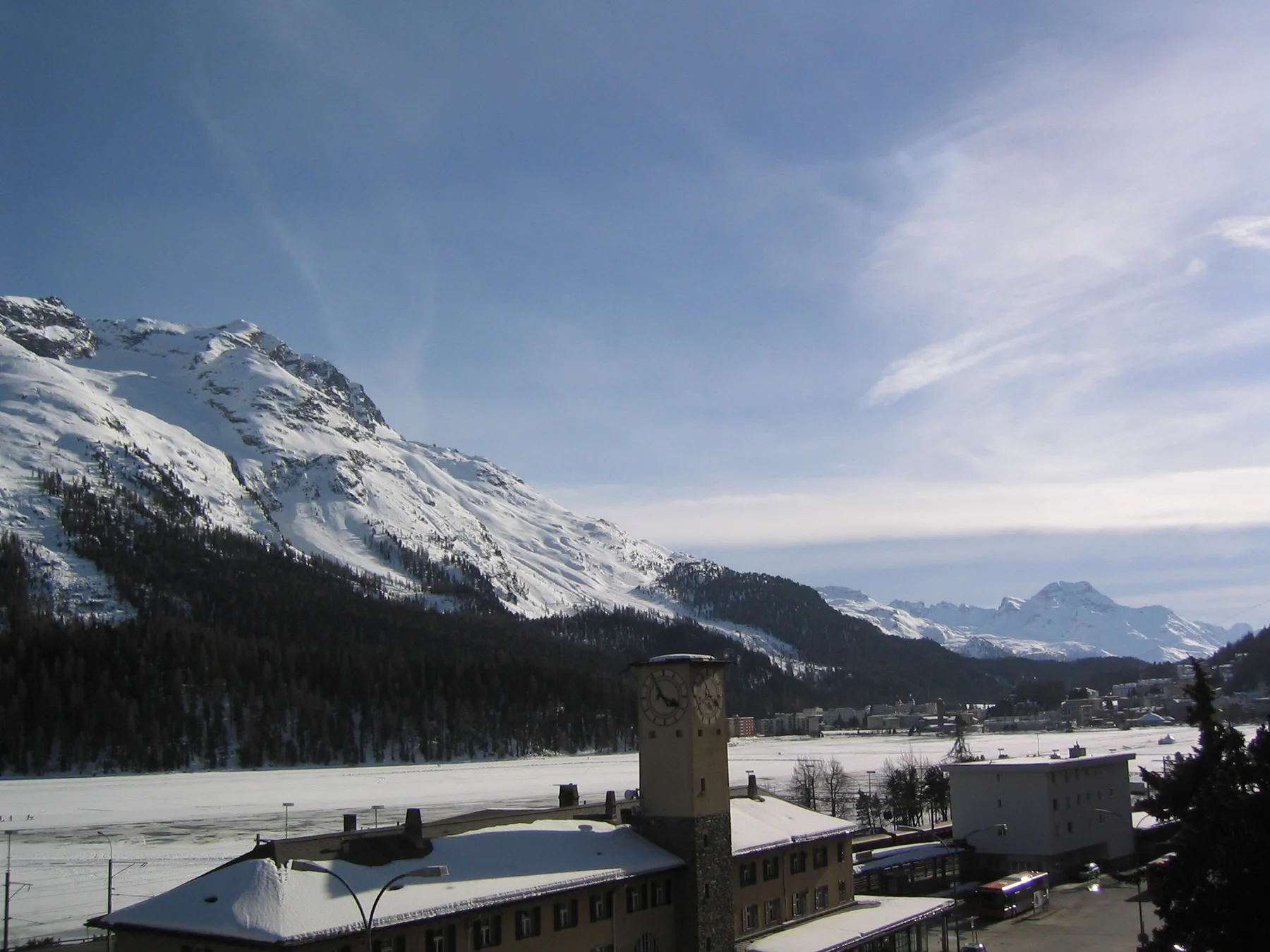 A station tower and snowy roofs sit below a mountain slope and wispy clouds.