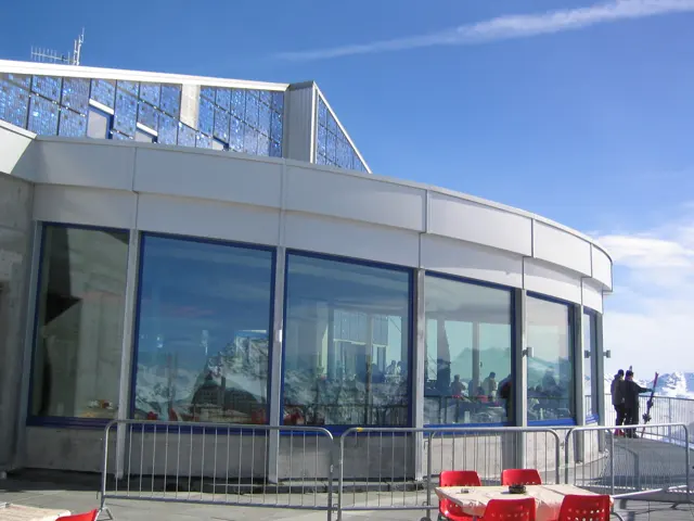 A rounded glass-fronted building reflects snowy mountains beside red terrace chairs.