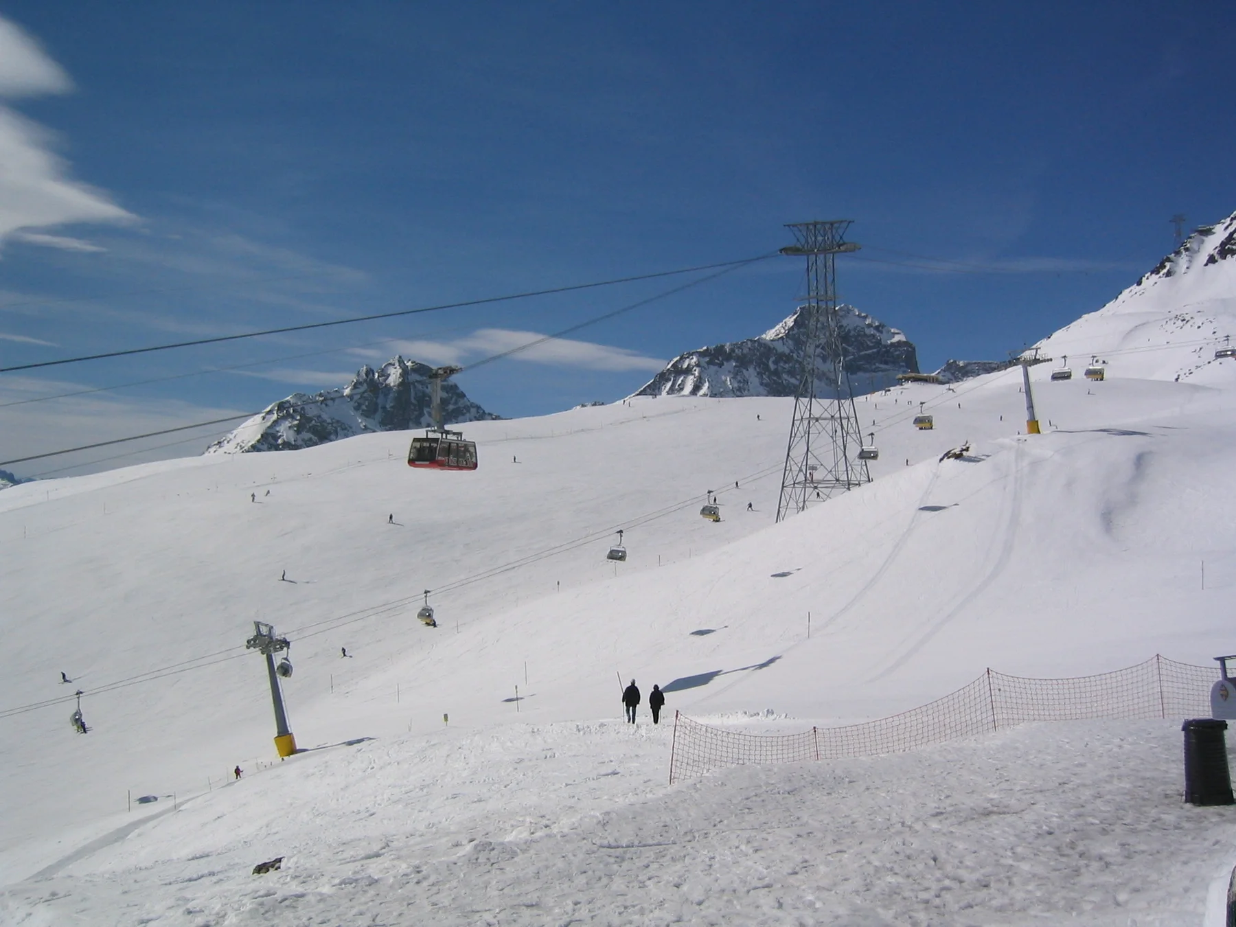 Cable towers and suspended cabins cross a high snowy slope above small figures.
