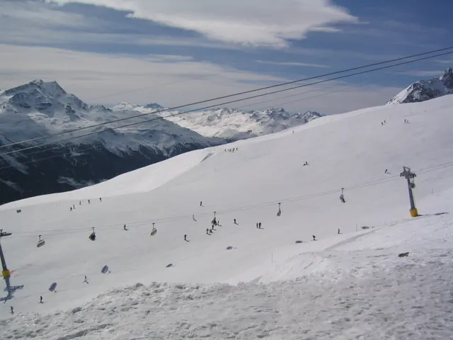 Small skiers traverse a white slope beneath overhead cables and distant peaks.