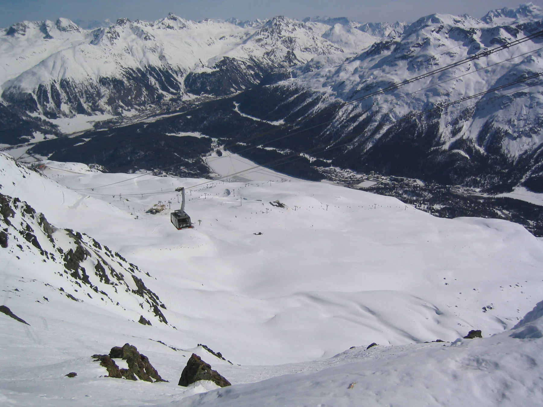 A gondola hangs above a steep snow basin overlooking a distant mountain valley.