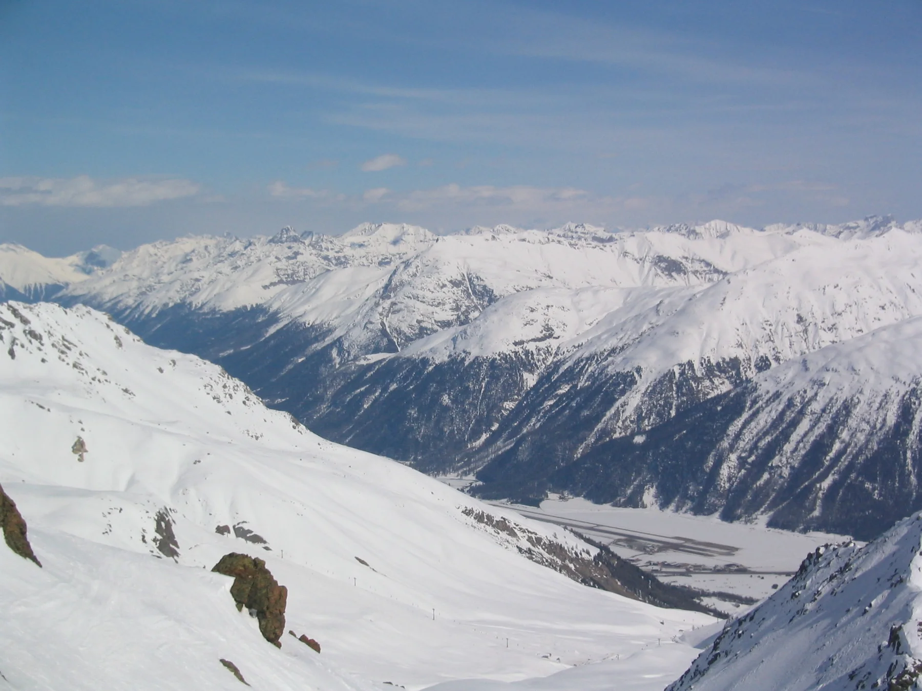 Smooth snow slopes with exposed rocks lead toward a long white mountain range.