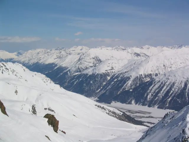 Smooth snow slopes with exposed rocks lead toward a long white mountain range.